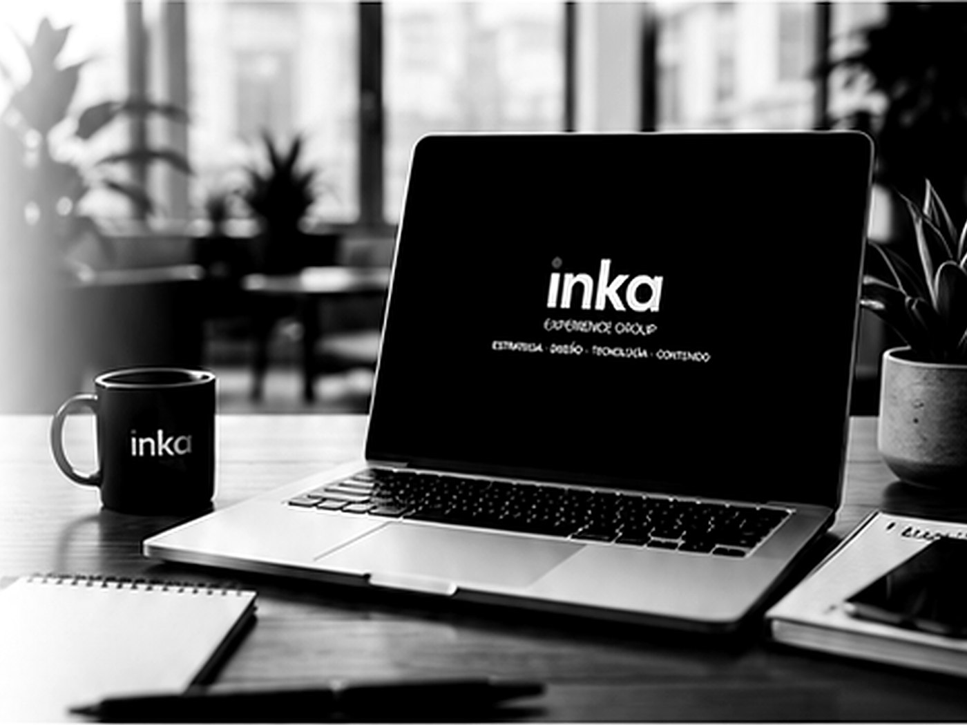 Laptop sobre un escritorio de oficina mostrando la pantalla de bienvenida de Inka Experience Group, junto a una taza de café de la marca, foto en blanco y negro